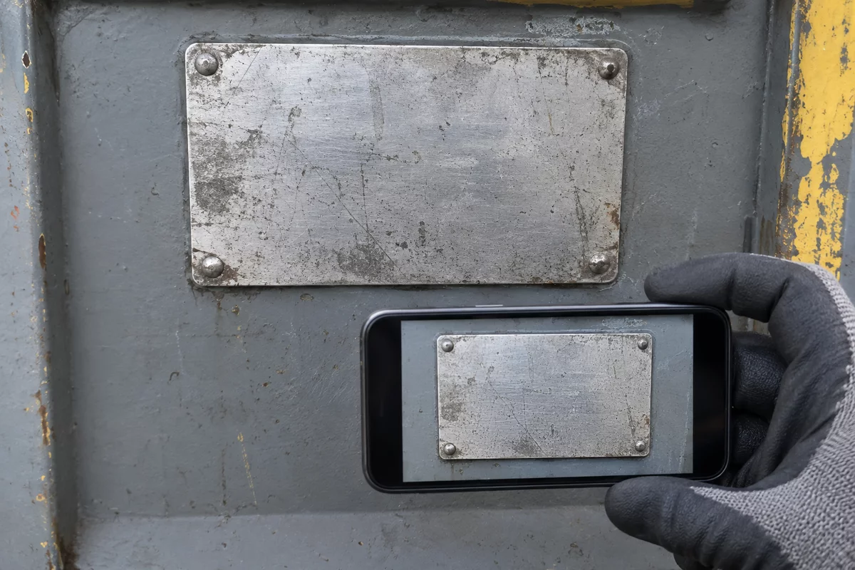 Illustration of a gloved hand framing one blank machine data plate with a smartphone, whose screen shows the same plate.